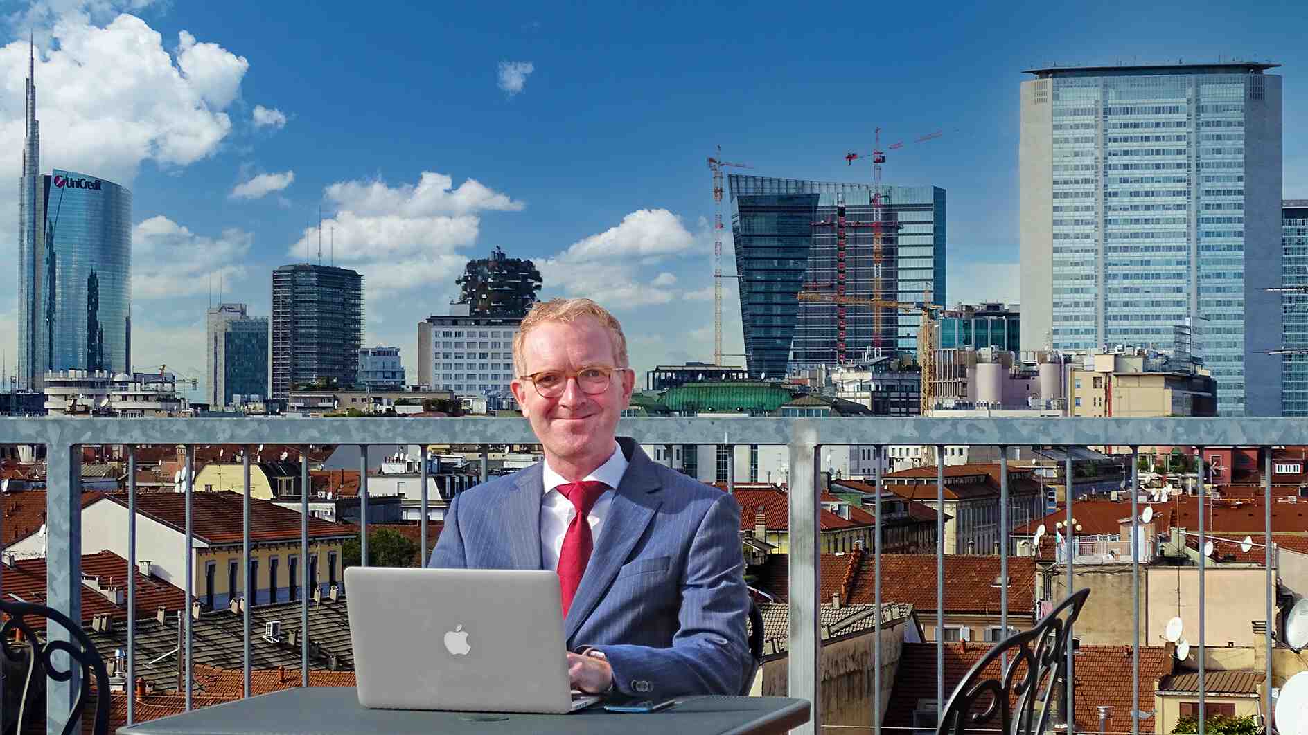 Man in suit and tie working on a MacBook on a rooftop in front of the skyline of Milan
